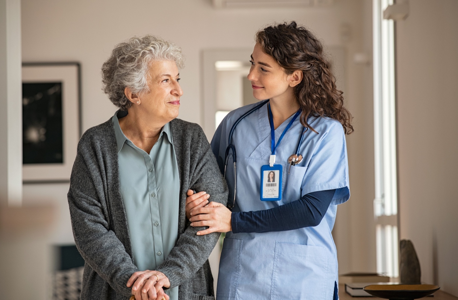 A caregiver and a senior woman in assisted living linking arms while smiling at each other.