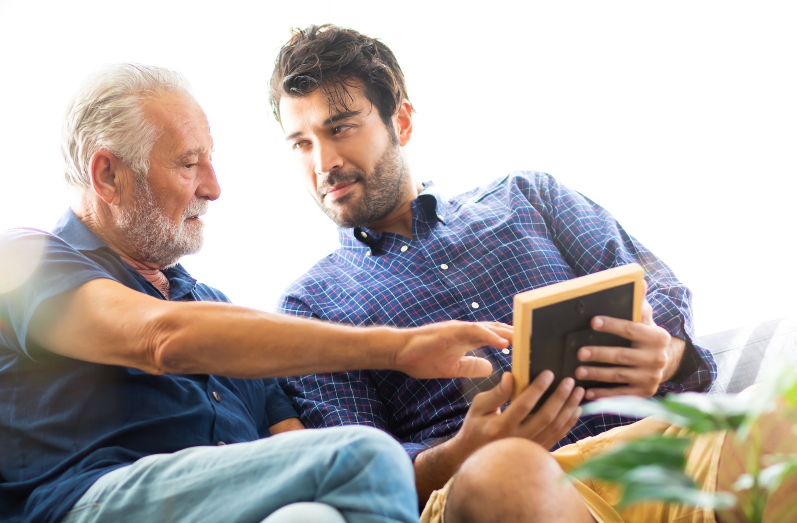 Adult son and older father reminiscing over a photograph on a couch.