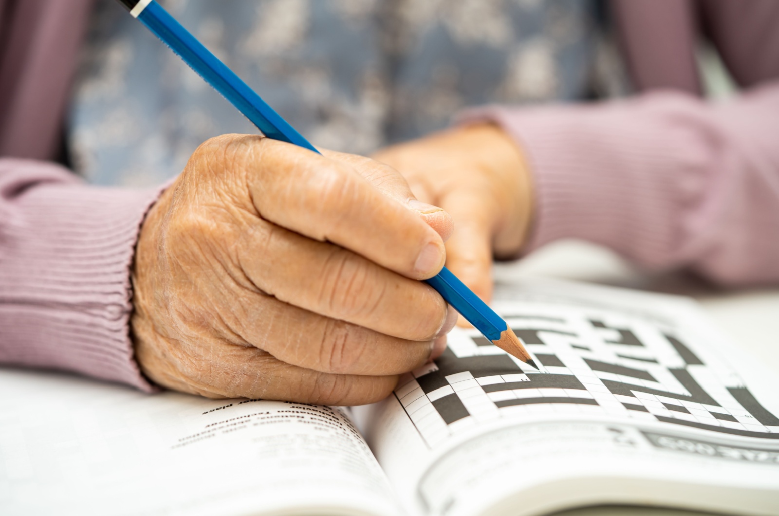 A close-up image of a senior holding a blue pencil in their right hand and doing a crossword puzzle.