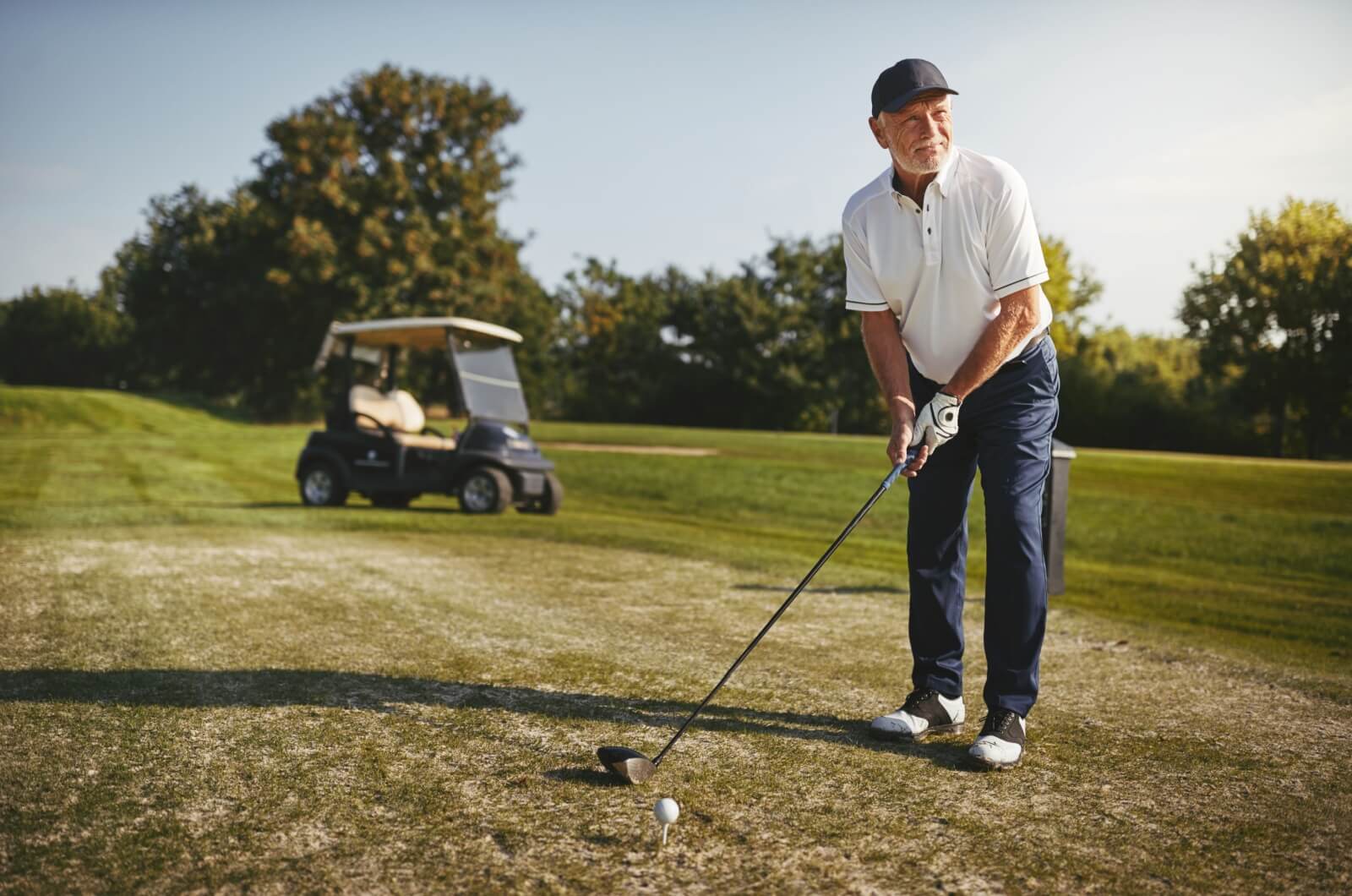 An older adult ready to take a shot with their golf club on a course with a golf cart in the background.
