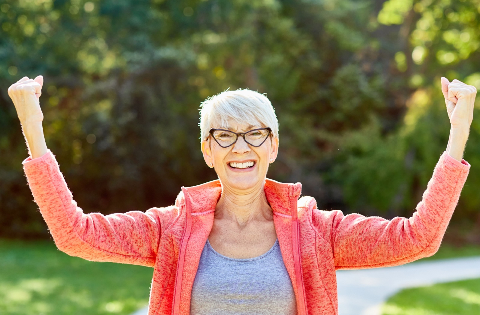 An older adult smiling and flexing their arms while on a walk outdoors in a park.