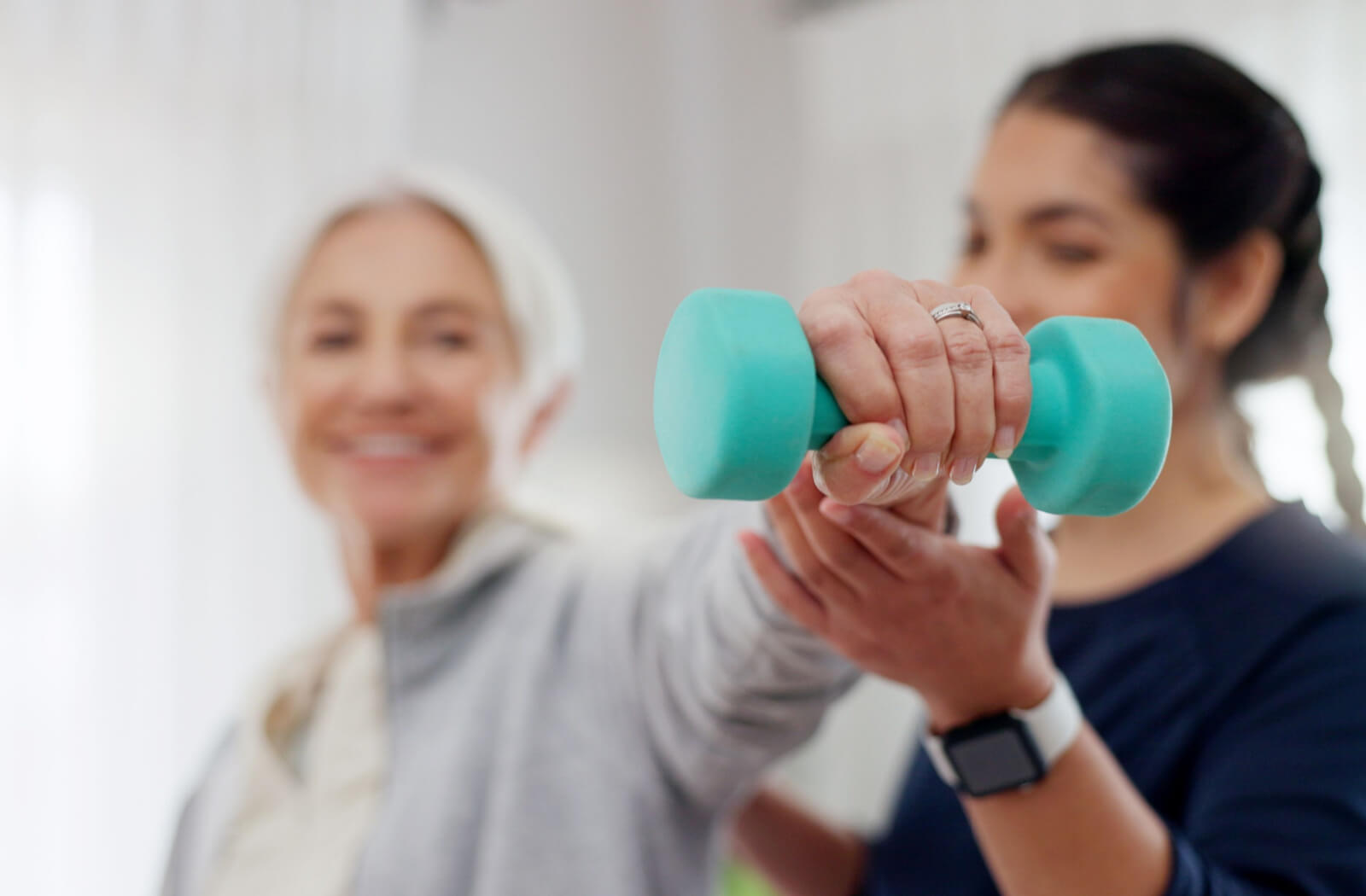 A caregiver helps a smiling older adult lift a small blue weight as a strength exercise