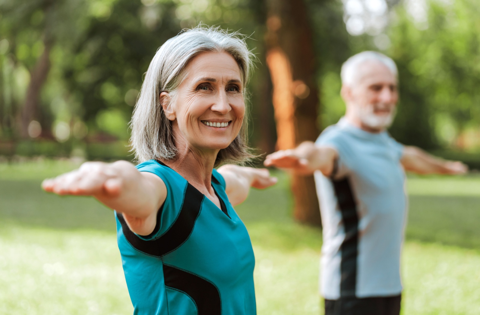 Two older adults doing Tai Chi in a park, smiling in the sun.