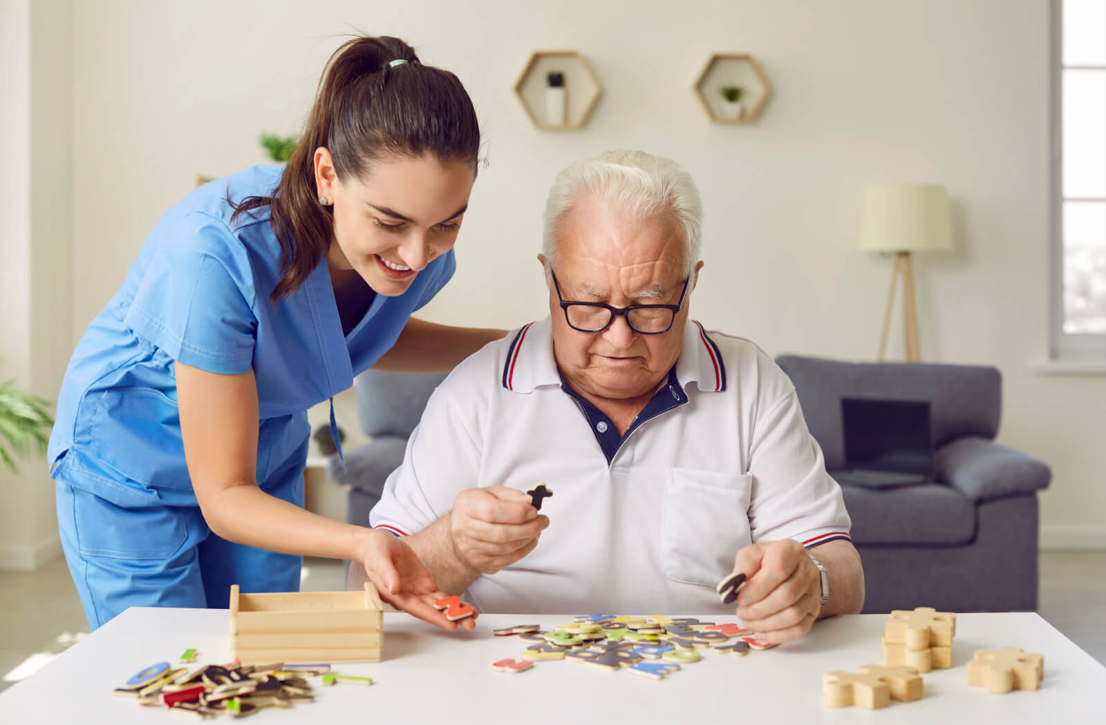 A smiling caregiver helps an older adult lay out wooden letters during a brain-strengthening exercise in their living room in memory care