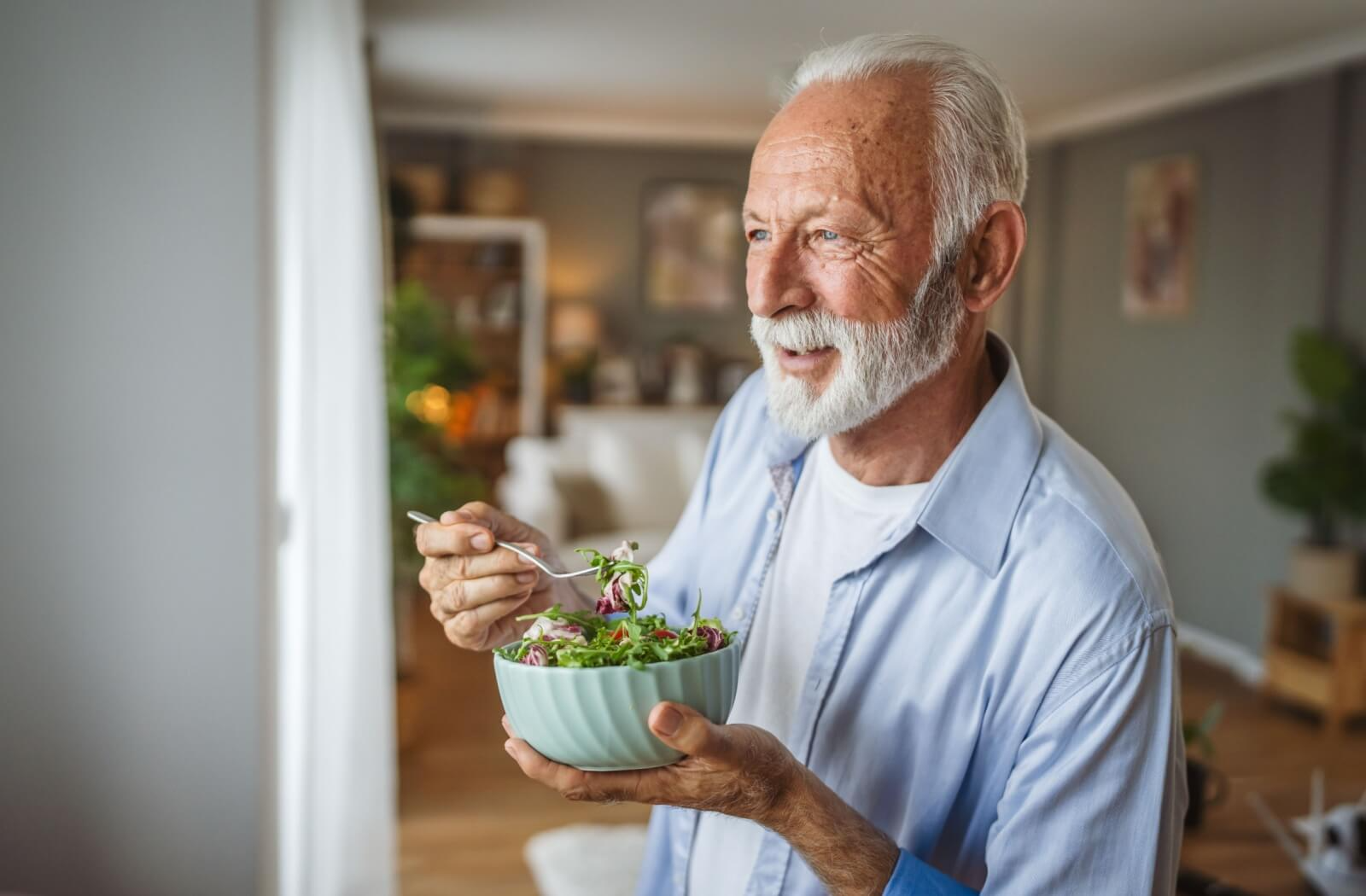 A senior eats a salad for a snack, with nuts for extra protein, while looking out the window of their senior living home