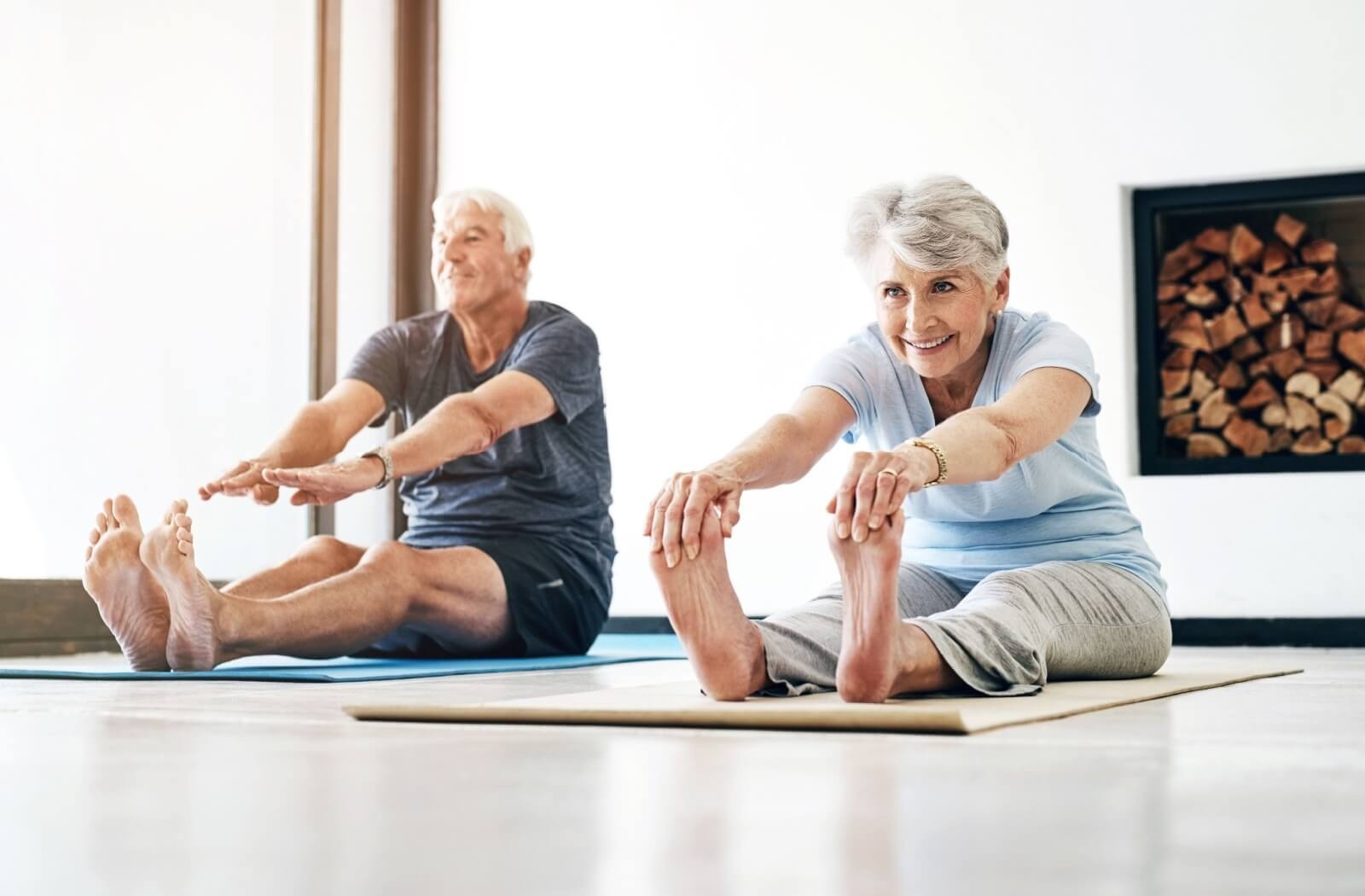 Two older adults sit on the floow and reach forward to touch their days while stretching in the morning

