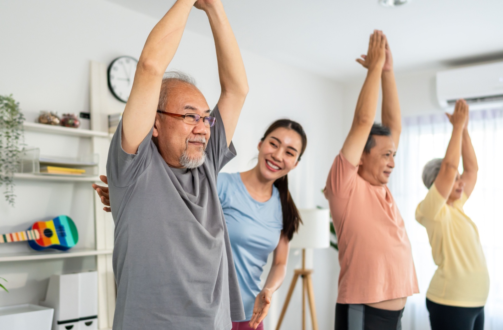 A caregiver helps three older adults extend their arms overhead and stretch during a morning fitness program in senior living